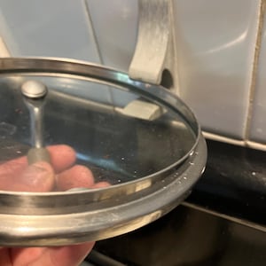 May include: A stainless steel and glass lid for a pot or pan. The lid is being held near a wall-mounted, brushed metal pot lid holder. The holder is mounted on white square tiles.
