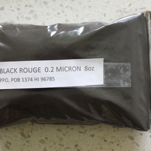 Könnte beinhalten: Ein durchsichtiger Plastikbeutel gefüllt mit schwarzem Rouge-Pulver. Das Etikett lautet "BLACK ROUGE 0.2 MICRON 8oz PPO, POB 1374 HI 96785".