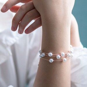 May include: A delicate bracelet featuring three strands of faux pearls on a thin, rose-gold colored chain. The bracelet is worn on a wrist, with a white shirt and a blue background. The pearls are round and evenly spaced.