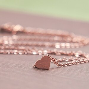 K&ouml;nnte beinhalten: Eine Halskette mit Herzanh&auml;nger in Ros&eacute;gold. Die filigrane Kette tr&auml;gt einen kleinen, massiven Herzanh&auml;nger. Die Halskette wird auf einer Holzoberfl&auml;che mit unscharfem Hintergrund pr&auml;sentiert. Der Schmuck ist ein romantisches Accessoire.