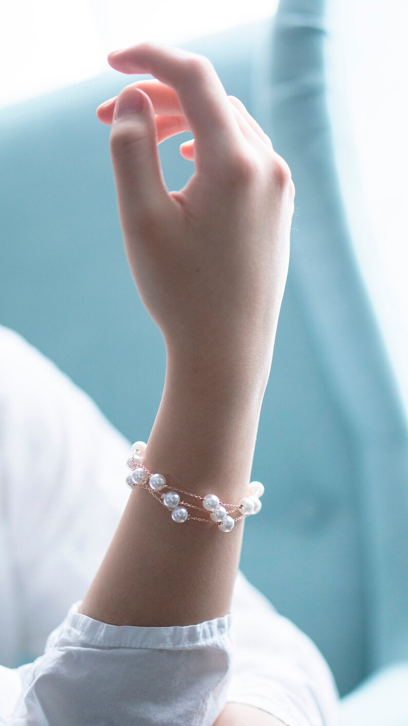 May include: A delicate bracelet featuring multiple strands of small, white pearls. The bracelet is strung on a thin, rose-gold colored chain. The image shows the bracelet on a person's wrist, with a white shirt in the background.