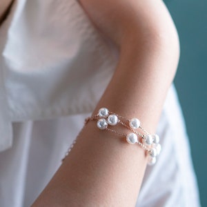 May include: A delicate bracelet featuring multiple strands of white pearls. The bracelet is set on a gold-toned chain and is worn on an arm, against a backdrop of a white shirt and a teal background. The jewelry is elegant and suitable for special occasions.
