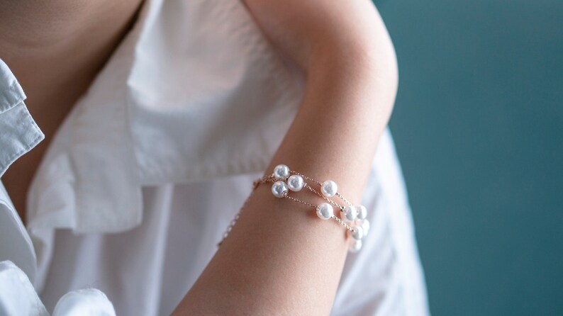 May include: A delicate bracelet featuring multiple strands of white pearls. The bracelet is set on a gold-toned chain and is worn on an arm, against a backdrop of a white shirt and a teal background. The jewelry is elegant and suitable for special occasions.