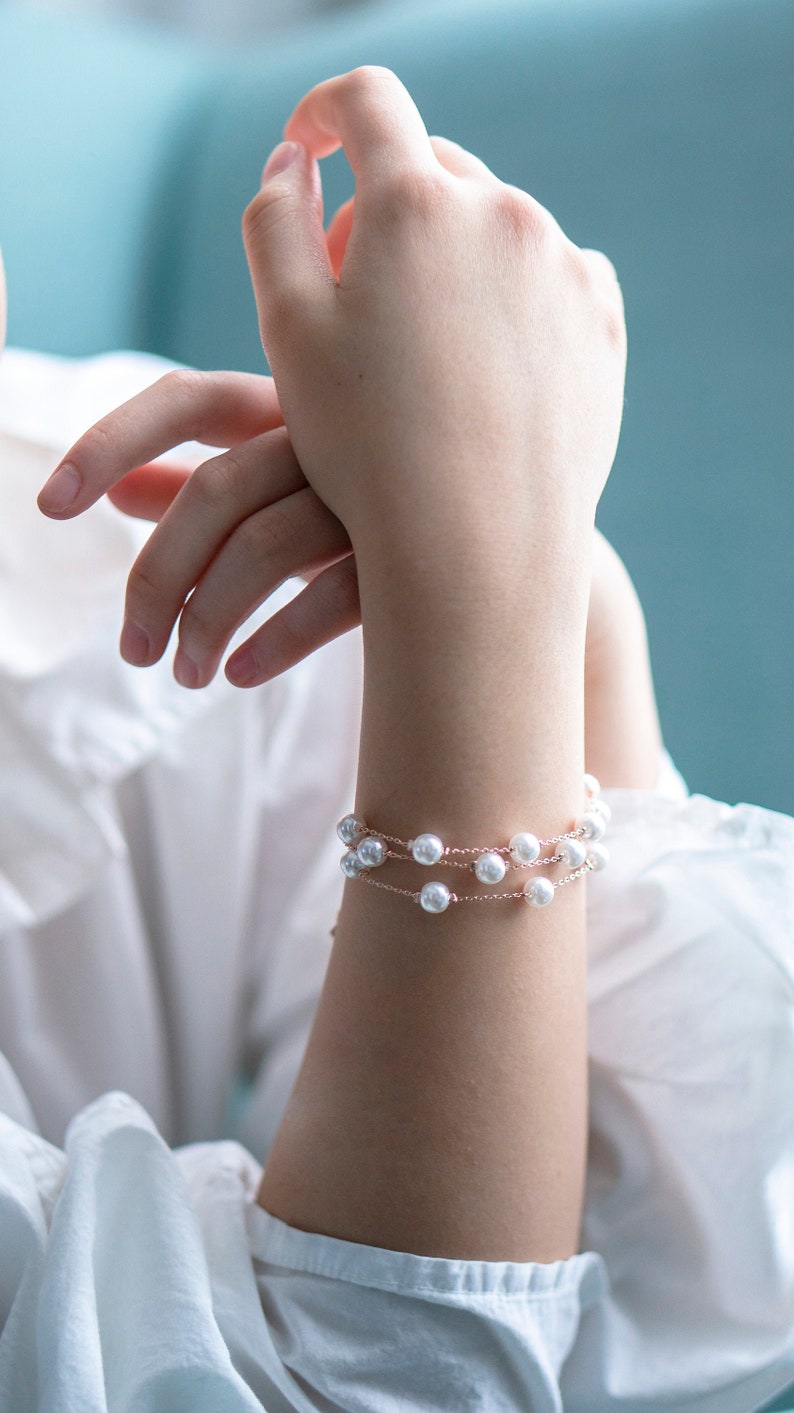 May include: A delicate bracelet featuring three strands of faux pearls on a thin, rose-gold colored chain. The bracelet is worn on a wrist, with a white shirt and a blue background. The pearls are round and evenly spaced.