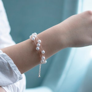 May include: A delicate, double-strand pearl bracelet. The bracelet features small, round, white pearls strung on a thin, gold-colored chain. The bracelet is worn on a person's wrist, with a white shirt in the background.