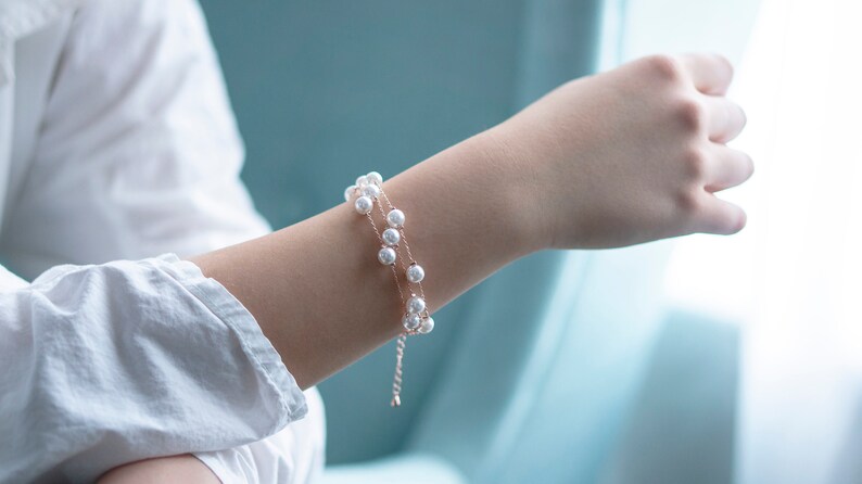 May include: A delicate, double-strand pearl bracelet. The bracelet features small, round, white pearls strung on a thin, gold-colored chain. The bracelet is worn on a person's wrist, with a white shirt in the background.