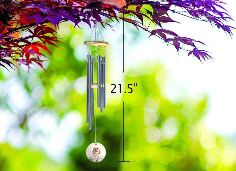 May include: A 21.5-inch wind chime with silver-colored tubes and a wooden top and bottom. A round, wooden pendant at the bottom features a dog portrait and the name "Lola". The chime hangs from a tree branch with purple leaves.
