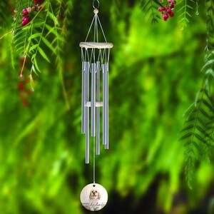 May include: Silver wind chimes with a wooden top and a round wooden pendant featuring a dog's portrait and the name "Lola". The chimes hang against a blurred green background with red berries.