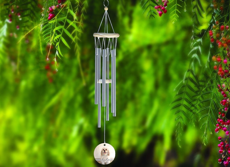 May include: Silver wind chimes with a wooden top and a round wooden pendant featuring a dog's portrait and the name "Lola". The chimes hang against a blurred green background with red berries.