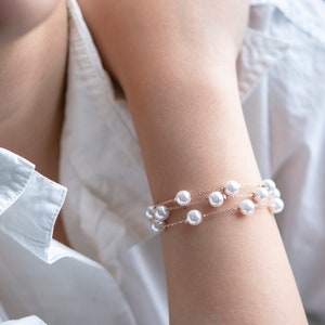 May include: A delicate bracelet featuring three strands of white pearls strung on a fine, rose-gold chain. The bracelet is displayed on a person's arm, against the backdrop of a white shirt.