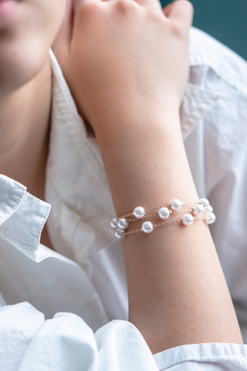 May include: A delicate bracelet featuring three strands of white pearls strung on a fine, rose-gold chain. The bracelet is displayed on a person's arm, against the backdrop of a white shirt.