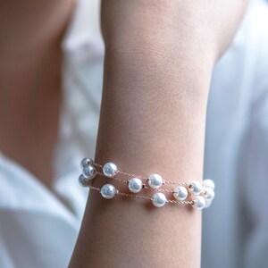 May include: A delicate bracelet featuring three strands of white faux pearls strung on a fine, rose-gold colored chain. The bracelet is wrapped around a wrist, showcasing the jewelry's elegant design. The pearls are evenly spaced.