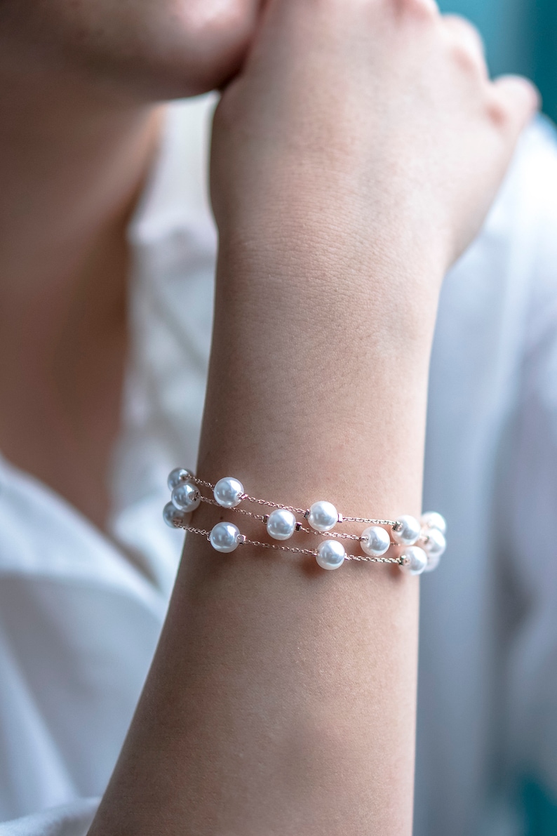 May include: A delicate bracelet featuring three strands of white faux pearls strung on a fine, rose-gold colored chain. The bracelet is wrapped around a wrist, showcasing the jewelry's elegant design. The pearls are evenly spaced.
