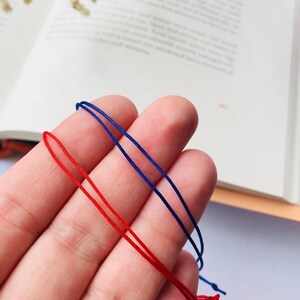 Peut inclure: Deux cordons de ficelle, l'un rouge et l'autre bleu, sont tenus dans une main. Les cordons sont fins et semblent être en coton.