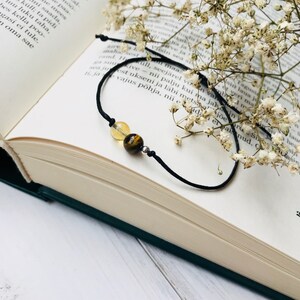 Puede incluir: Una pulsera de cordón negro con una cuenta de citrino, una cuenta de ojo de tigre y una pequeña cuenta de plata. La pulsera se muestra en un libro abierto con flores secas.