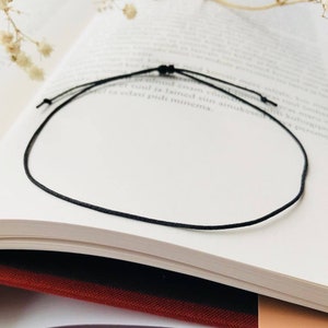 Puede incluir: Un collar sencillo de cordón negro, anudado, sobre un libro blanco.