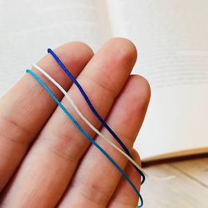 Puede incluir: Tres hebras de cordón en colores azul, blanco y azul claro. Los cordones se sostienen en una mano.