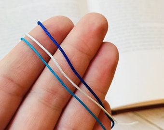 May include: Three strands of cord in blue, white, and light blue colors. The cords are held in a hand.