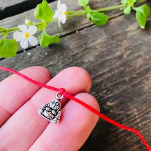 May include: A red string bracelet with a silver charm of a sitting Buddha.