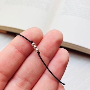 May include: A thin black cord bracelet featuring three small, polished silver-coloured spherical beads centred along the string. The cord is smooth and dark, providing a stark contrast to the reflective, metallic finish of the three round beads.