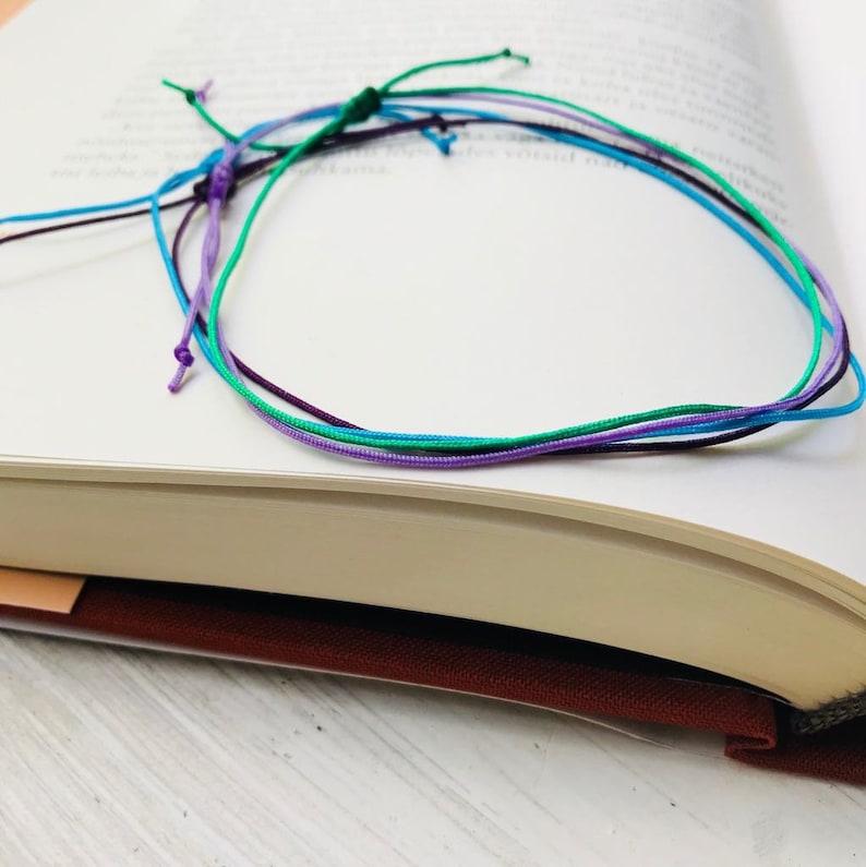 Peut inclure: Un ensemble de quatre bracelets d'amiti&eacute; color&eacute;s dans des tons de violet, bleu et vert. Les bracelets sont faits de cordons fins tress&eacute;s et sont nou&eacute;s ensemble.
