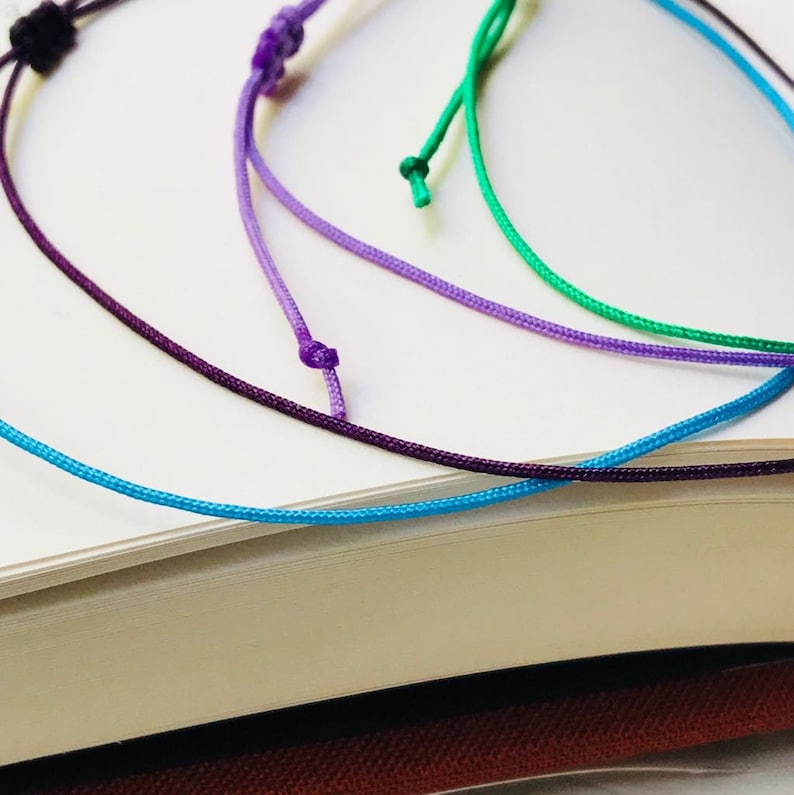 Peut inclure: Cinq brins de ficelle color&eacute;e en bleu, violet et vert sont nou&eacute;s ensemble. Les ficelles sont pos&eacute;es sur une surface blanche.