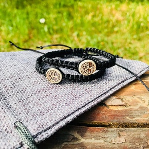 May include: Two black braided bracelets with wooden tree of life medallions. The bracelets are on a gray fabric surface, with a wooden table and green grass in the background. The bracelets are adjustable.