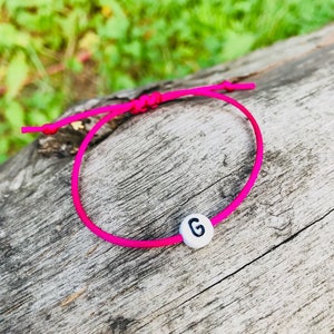May include: A vibrant pink bracelet with a white bead displaying the letter "G" in black. The bracelet is crafted from a thin, flexible cord and is knotted at the ends. The backdrop is a weathered wooden surface.