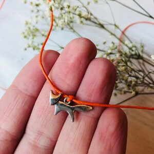 Puede incluir: Un charm de zorro plateado en una cuerda naranja. El charm es una silueta simple de un zorro.