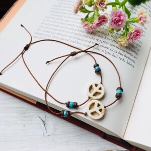 May include: Two adjustable brown cord bracelets featuring a circular white peace sign charm. Each bracelet is accented with small dark brown wooden beads and bright turquoise cylindrical beads, creating a simple and symmetrical design.