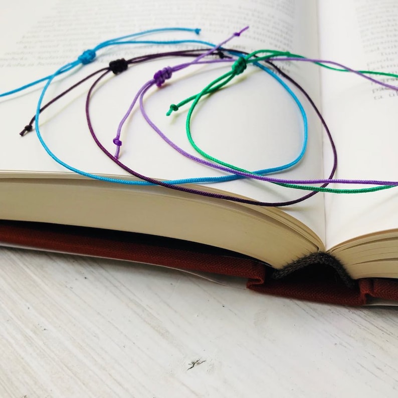 Peut inclure: Un ensemble de bracelets en corde de diff&eacute;rentes couleurs, dans des tons de bleu, violet et vert. Les bracelets sont li&eacute;s ensemble et dispos&eacute;s sur un livre.