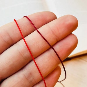 Peut inclure: Deux brins de cordon rouge, l'un d'une nuance de rouge plus foncée que l'autre, sont tenus dans la main d'une personne. Les cordons sont lisses et semblent être faits d'une fibre naturelle.