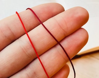 May include: Two strands of red cord, one a darker shade of red than the other, are held in a person's hand. The cords are smooth and appear to be made of a natural fiber.
