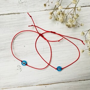 May include: Two red string bracelets, each featuring a small, round, blue evil eye charm. The bracelets are displayed on a white wooden surface, with dried white flowers in the background. The string is knotted for adjustability.