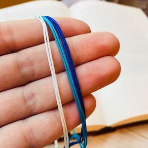 May include: Four strands of cord in different shades of blue and white. The cords are held in a hand.