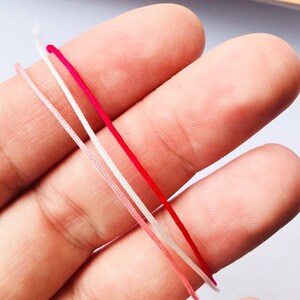 May include: A close-up shot of four colourful cords: pale pink, white, red, and a deeper pink. The cords are thin and appear to be made of a smooth material. The cords are held in a person's hand, with the focus on the colourful strands.
