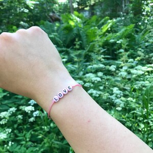 May include: A pink string bracelet featuring four round, light pink beads with the black letters L, O, V, and E printed on them, spelling out the word LOVE. The bracelet is thin and simple, designed to be worn around the wrist as an accessory.