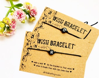 Puede incluir: Dos pulseras de deseos con hilo negro y cuentas de letras, una con "D" y otra con "R". Las pulseras están en tarjetas marrones con las palabras "WISH BRACELET" impresas. Las tarjetas también tienen el texto "pide un deseo, ata la pulsera a tu muñeca, cuando se rompa tu deseo puede hacerse realidad."