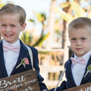 May include: Two young boys in navy blue suits with pink bow ties and boutonnieres. Each boy holds a wooden sign with white text. The sign on the left reads "Brett here comes your..." and the sign on the right reads "...seen".