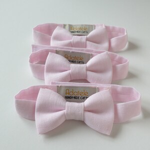 May include: Four light pink headbands with bow accents are arranged on a white surface. Each headband features a bow in the center and a rectangular tag with the text "ADAT&Eacute;LE HANDMADE GIFTS". The headbands are made of a soft fabric.