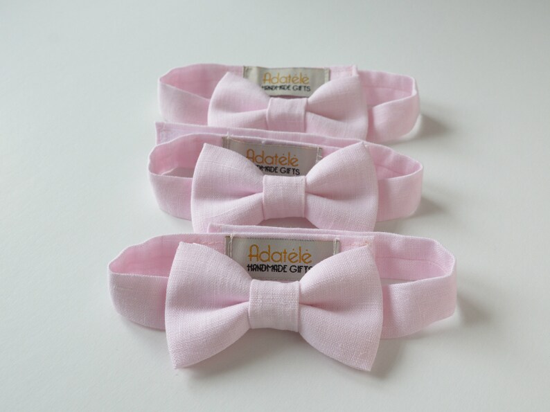 May include: Four light pink headbands with bow accents are arranged on a white surface. Each headband features a bow in the center and a rectangular tag with the text "ADAT&Eacute;LE HANDMADE GIFTS". The headbands are made of a soft fabric.