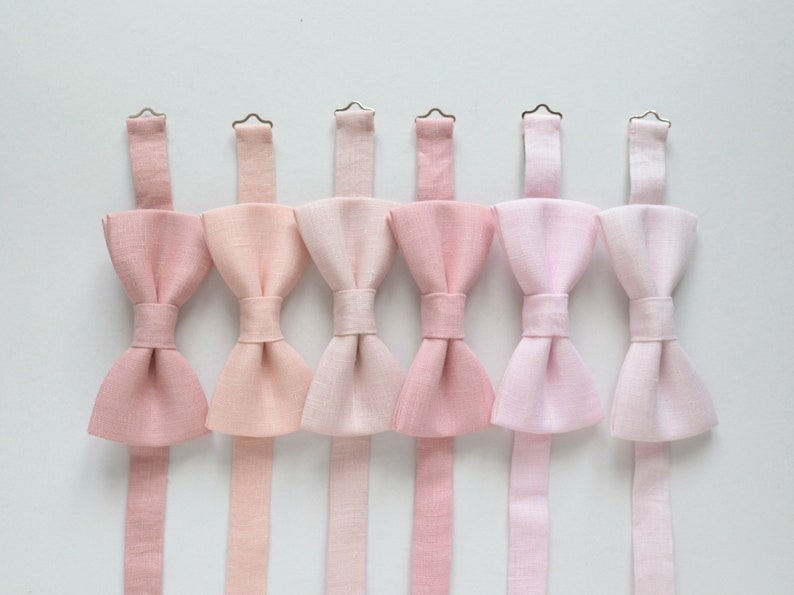 May include: Six pink bow ties in varying shades, arranged in a row. Each bow tie has a matching adjustable neck strap and a metal clasp. The bow ties are made of a textured fabric, and the background is plain white.
