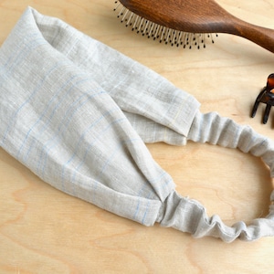 Può includere: Un cerchietto in lino grigio chiaro con un sottile motivo a quadri blu e bianco. Il cerchietto è elastico e può essere indossato in vari modi.