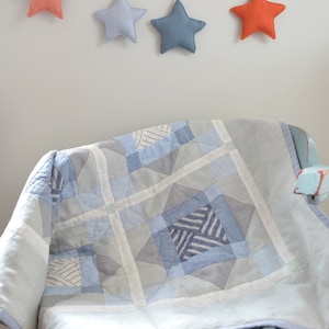 Puede incluir: Una colcha de retazos de color azul claro y gris con un diseño de estrellas está drapeada sobre una silla de madera azul claro. Una guirnalda de estrellas de tela de color naranja y azul cuelga sobre la silla.