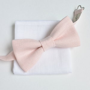 May include: A light pink bow tie and a white pocket square are displayed on a white background. The bow tie is made of a textured fabric and is neatly tied. The pocket square is square and made of a smooth fabric. The bow tie's strap is visible.