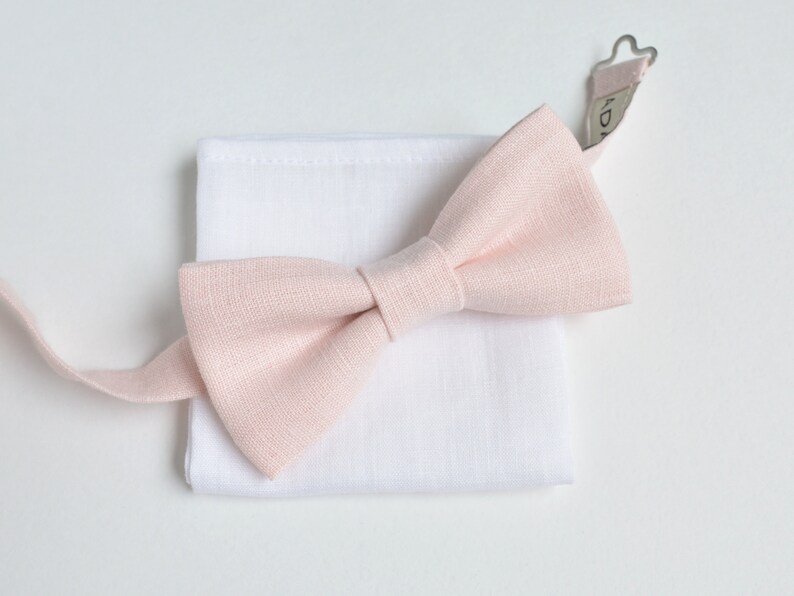 May include: A light pink bow tie and a white pocket square are displayed on a white background. The bow tie is made of a textured fabric and is neatly tied. The pocket square is square and made of a smooth fabric. The bow tie's strap is visible.