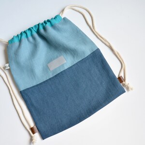 Può includere: Uno zaino con coulisse a due toni nei toni del blu e del verde acqua. La sezione superiore è azzurra, che sfuma in un blu più scuro nella parte inferiore. Presenta una piccola etichetta rettangolare e cinghie di corda color crema con dettagli in pelle.