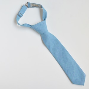 May include: A light blue necktie with a textured weave, featuring an adjustable neck strap. The tie is a solid color and appears to be made of a linen or cotton blend. The tag reads "Adelate, YOUR SIZE, YOUR STYLE."