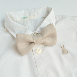 May include: A beige linen bow tie is centered on a white button-down shirt. The shirt has a small embroidered bunny near the bottom and a tag that reads "BENETTON BABY". The bow tie is made of a textured fabric.