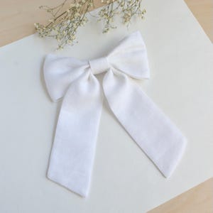 Puede incluir: Un lazo de lino blanco con largas colas fluidas. El lazo está centrado y atado, con las colas extendiéndose hacia abajo. Delicadas flores blancas están en el fondo. El lazo es probablemente para el cabello o como accesorio decorativo.